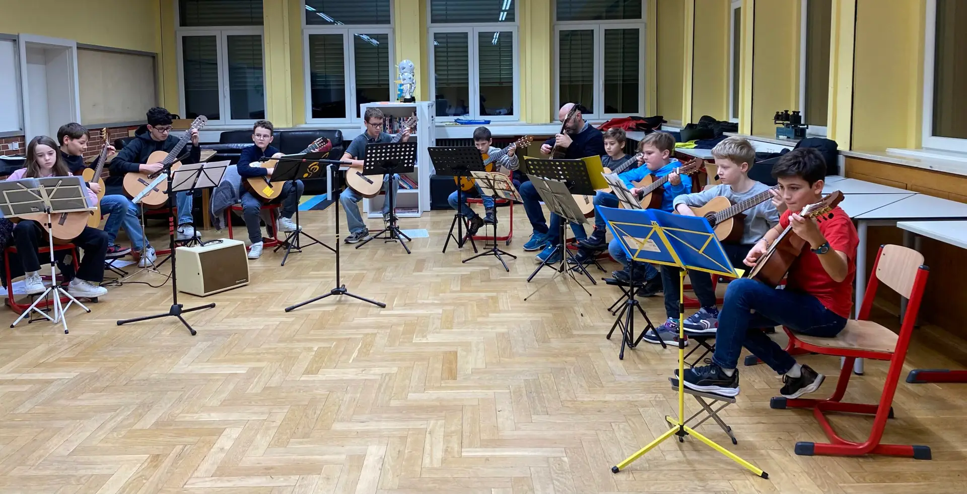 Gitarrenunterricht in großer Gruppe mit dem Musiklehrer und zahlreichen Kindern zwischen 6 und 9 Jahren.